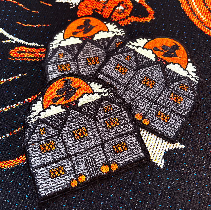 Three embroidered rectangular patches featuring gray haunted houses with orange windows, white clouds, and orange moon circles with black silhouettes, arranged on a dark patterned fabric background.