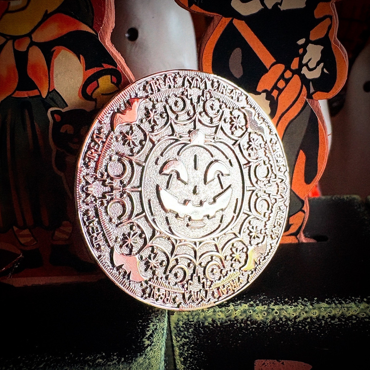 A round, ornate metallic stamp or medallion with a carved jack-o'-lantern face and floral scrollwork, shown in close-up against a dark, decorative background with Halloween-themed cutouts.