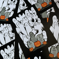 A close-up of overlapping rectangular prints or cards featuring a Halloween illustration: white ghostly hands rising from black ground, gray tombstones, and orange jack-o'-lantern pumpkins.