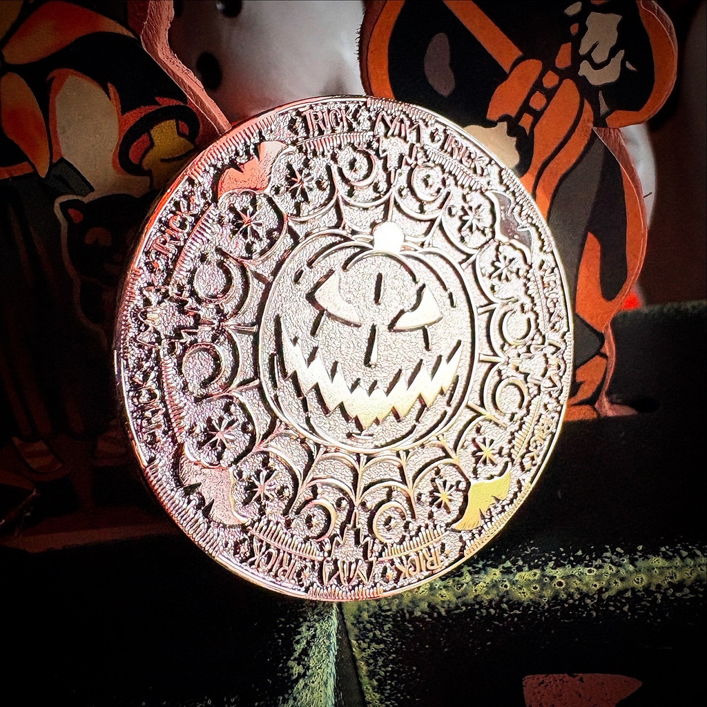 A round Halloween ornament featuring an intricately carved jack-o'-lantern face surrounded by floral and spiderweb motifs, with 'Happy Halloween' written around the edge. The ornament is shown hanging against a dark background with orange and black Halloween decorations.