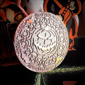 A round Halloween ornament featuring an intricately carved jack-o'-lantern face surrounded by floral and spiderweb motifs, with 'Happy Halloween' written around the edge. The ornament is shown hanging against a dark background with orange and black Halloween decorations.