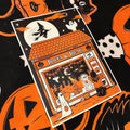 A Halloween-themed art print or card featuring an orange and black storefront with ghosts, pumpkins, a witch, and the words 'Tricks & Treats,' photographed on a black fabric background with orange pumpkin motifs.