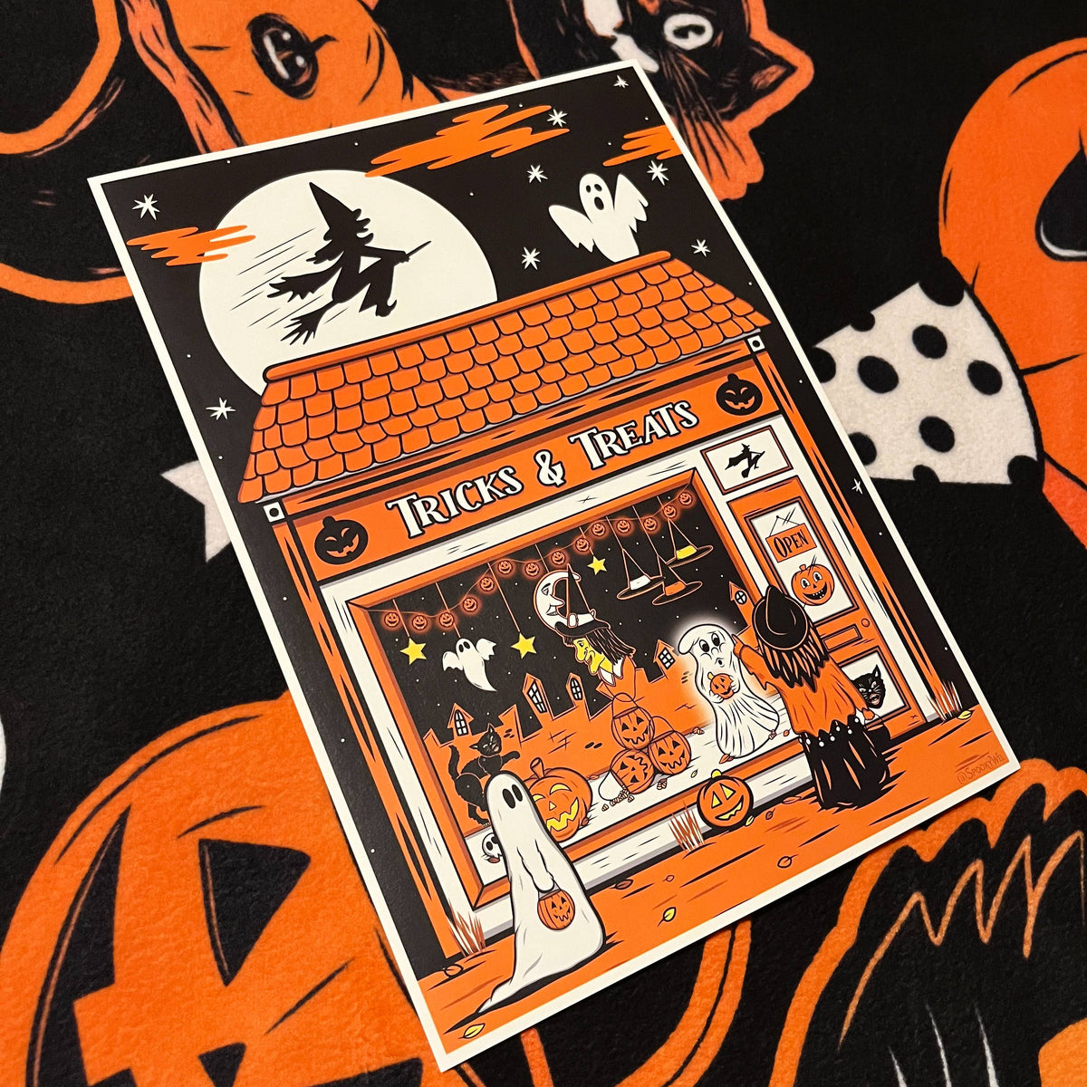 A Halloween-themed art print or card featuring an orange and black storefront with ghosts, pumpkins, a witch, and the words 'Tricks & Treats,' photographed on a black fabric background with orange pumpkin motifs.