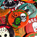 A close-up pile of round Halloween-themed pinback buttons showing assorted spooky designs, including a central orange button with a jack-o'-lantern, a green mask, a white skull, and the text 'It's Almost Time Kudos!'.