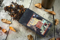 Halloween-Emporium Big Apple Chai black tea blend spilling from a matte black pouch on a rustic wooden table.