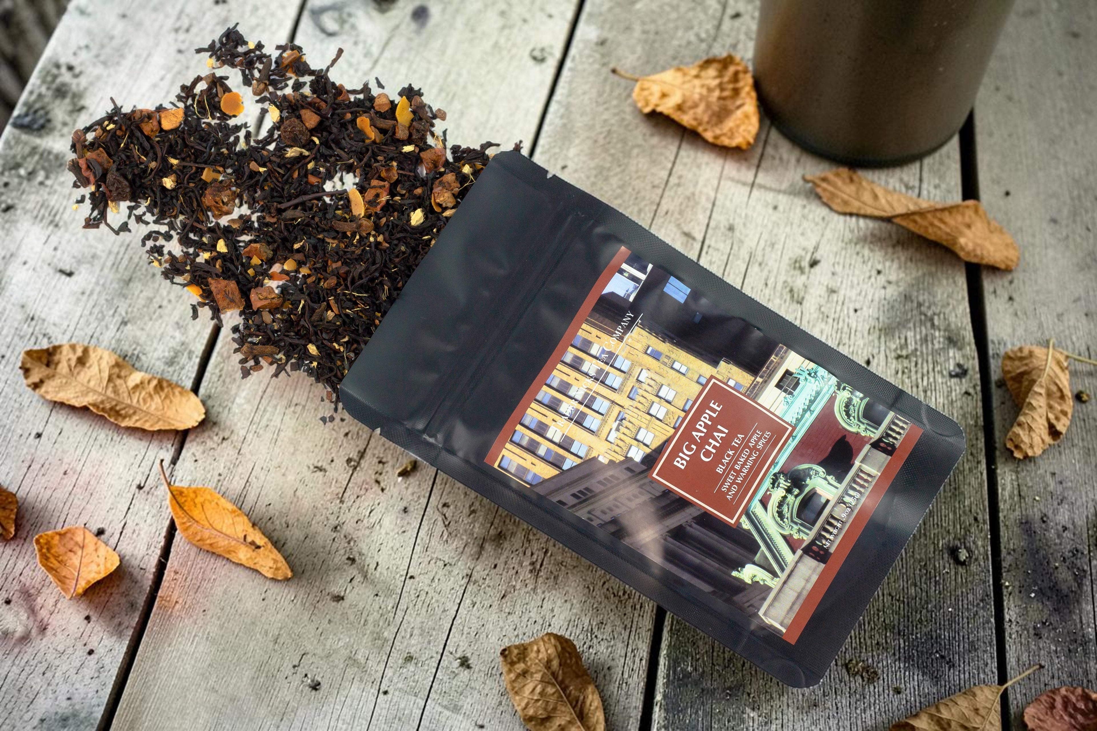Halloween-Emporium Big Apple Chai black tea blend spilling from a matte black pouch on a rustic wooden table.