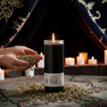 Halloween-Emporium 7 day black pillar candle in a glass jar, surrounded by scattered herbs for ritual practice.