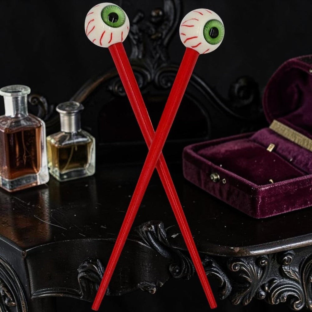 Halloween-Emporium pair of gothic hair sticks with realistic green eyeballs and blood-red shafts on a dark table.