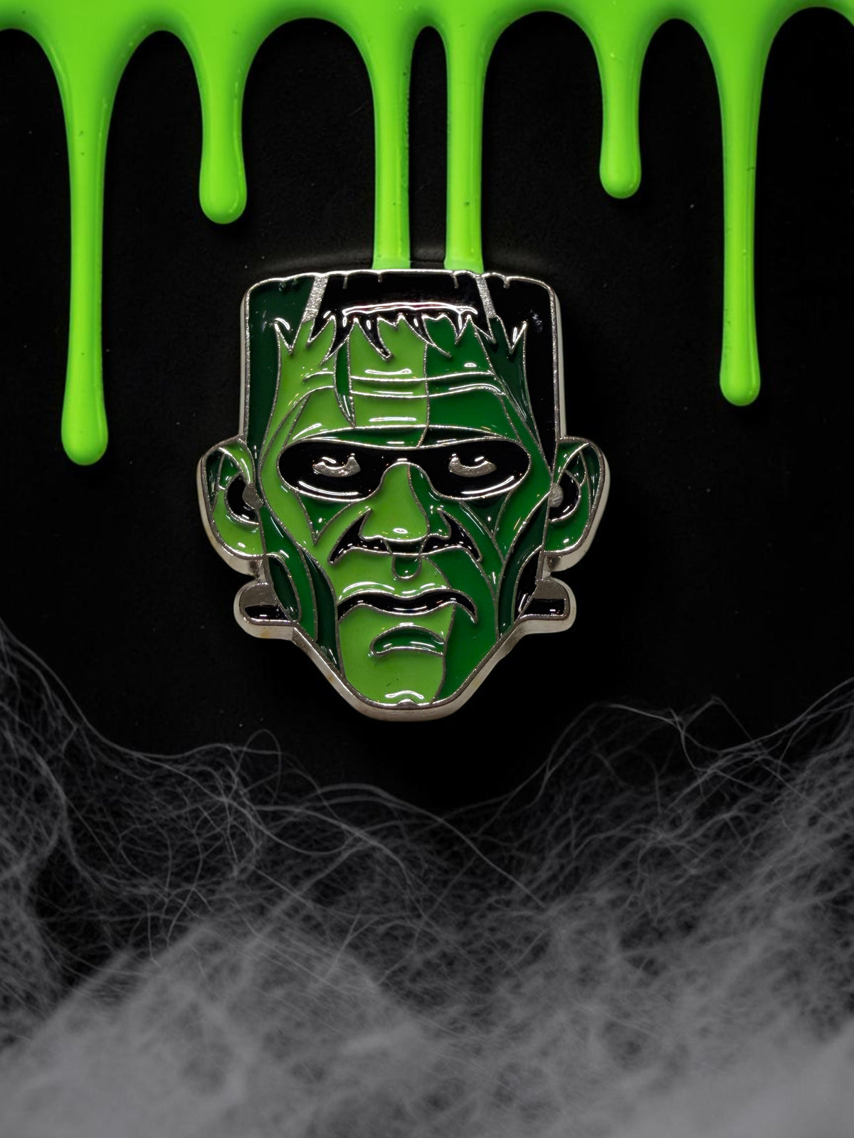 Halloween-Emporium Frankenstein's Monster enamel pin with green and black details on a spooky dripping background.