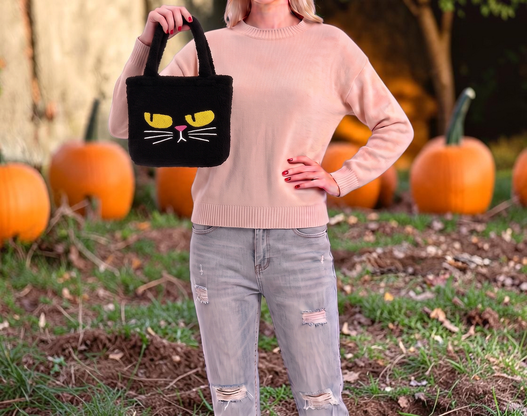 Halloween-Emporium furry black cat tote bag with a grumpy yellow-eyed expression held by a woman in a pumpkin patch.