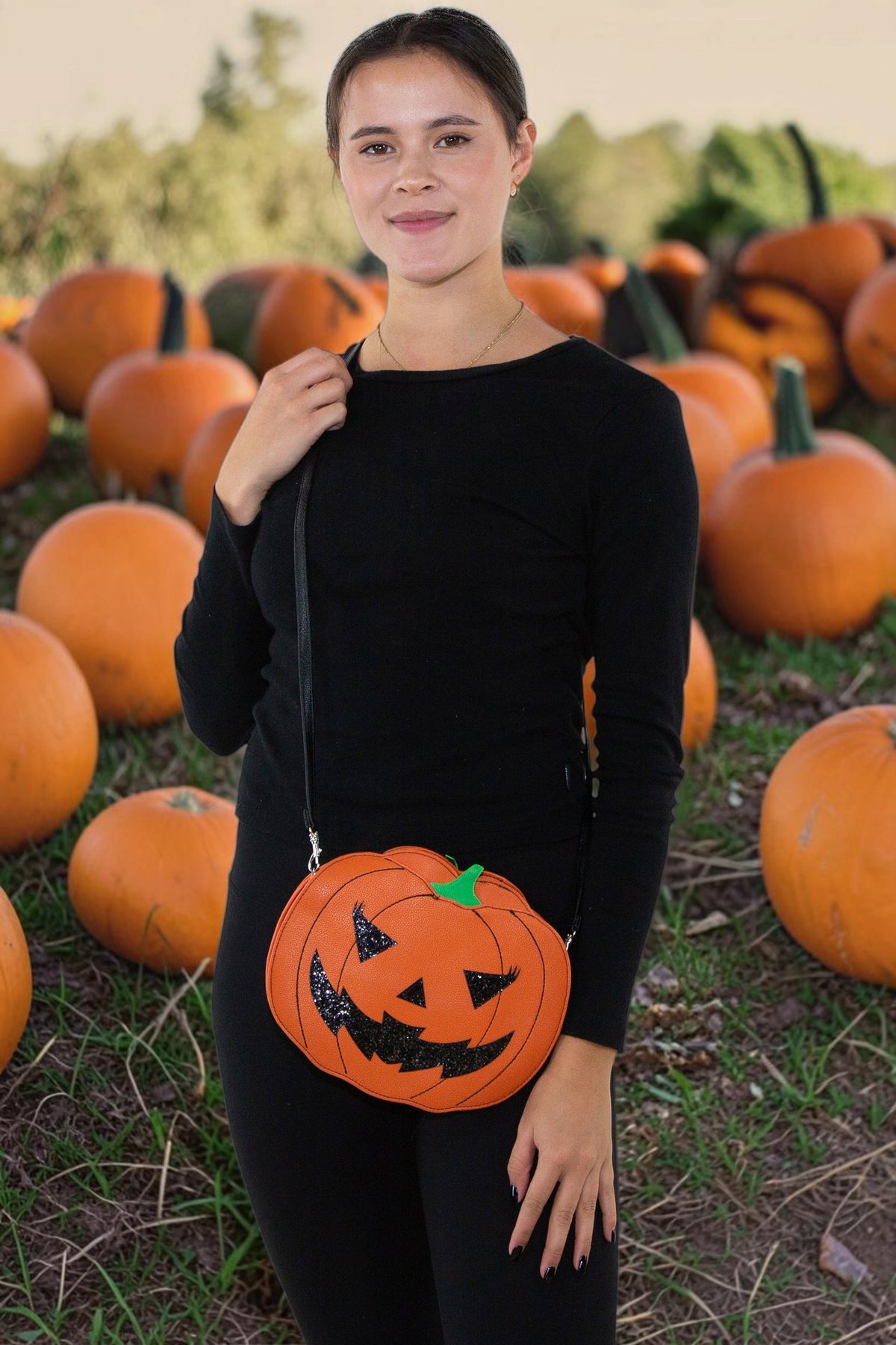 Halloween-Emporium glitter Jack O Lantern vinyl crossbody bag with orange strap worn by a woman in a pumpkin patch.