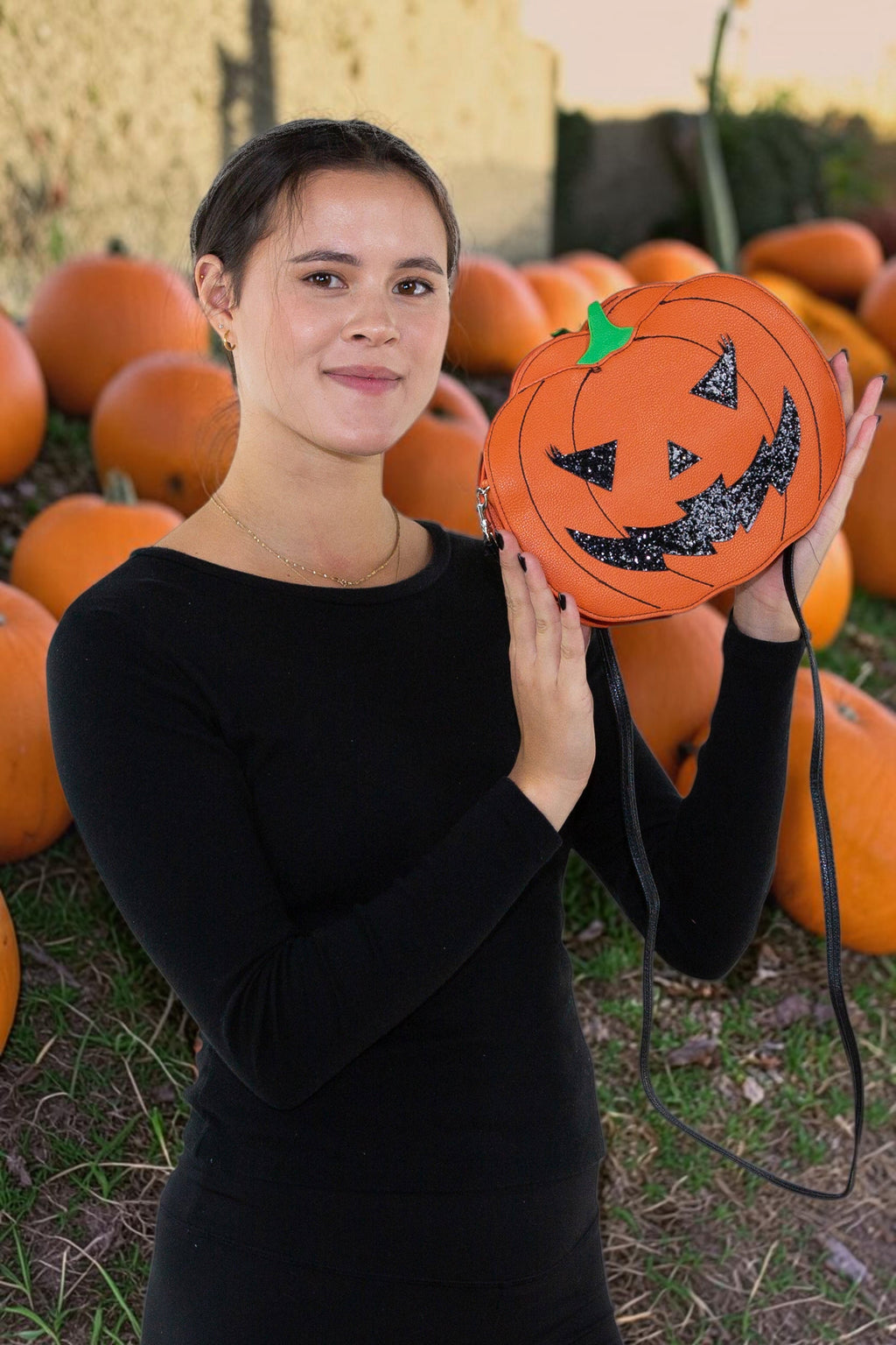 Halloween-Emporium glitter jack-o-lantern vinyl crossbody bag with orange strap held by a woman in a pumpkin patch.