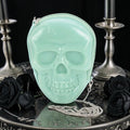 Halloween-Emporium glow-in-the-dark mint green vinyl skull crossbody bag with a silver chain strap on a dark table.