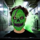 Halloween-Emporium neon green monster latex mask with skull fangs worn by a person in a smoky industrial setting.