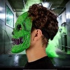 Halloween-Emporium neon green monster latex mask with skull fangs worn by a person in a smoky industrial setting.