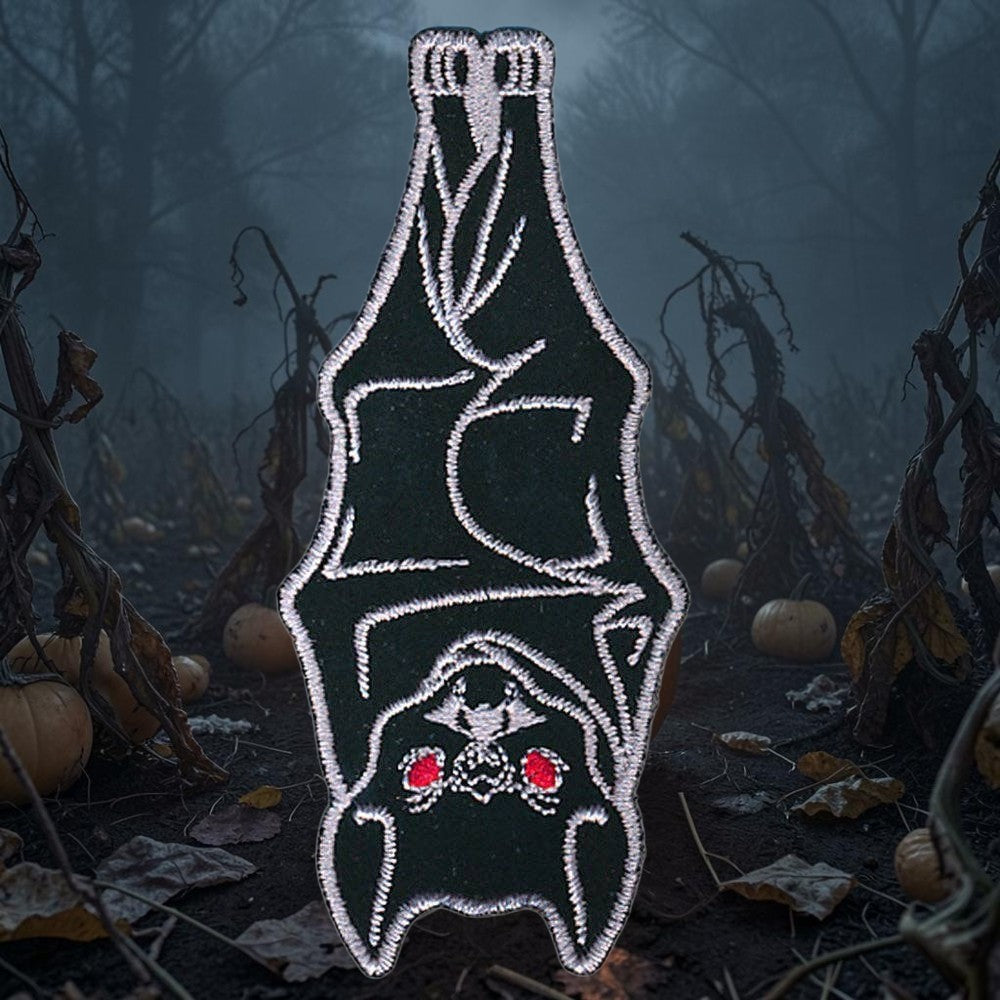 Halloween-Emporium hanging bat embroidered iron-on patch with white stitching and red eyes on a dark pumpkin patch