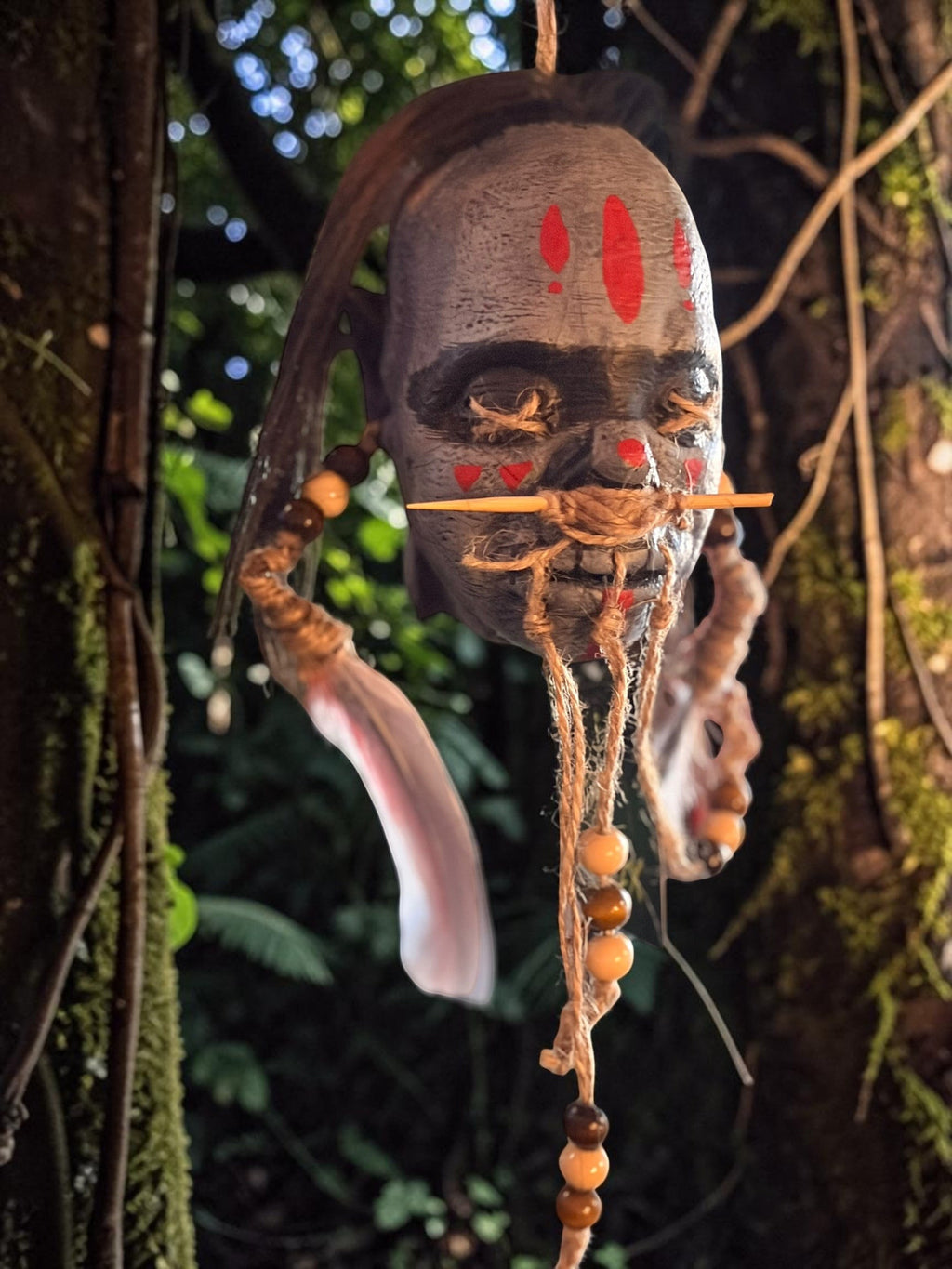 Halloween-Emporium latex shrunken head prop with red painted markings, hand-punched hair, and beaded feather accents.