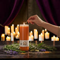 Halloween-Emporium 7 day orange candle in glass jar being lit by a hand with herbs on a dark wooden table