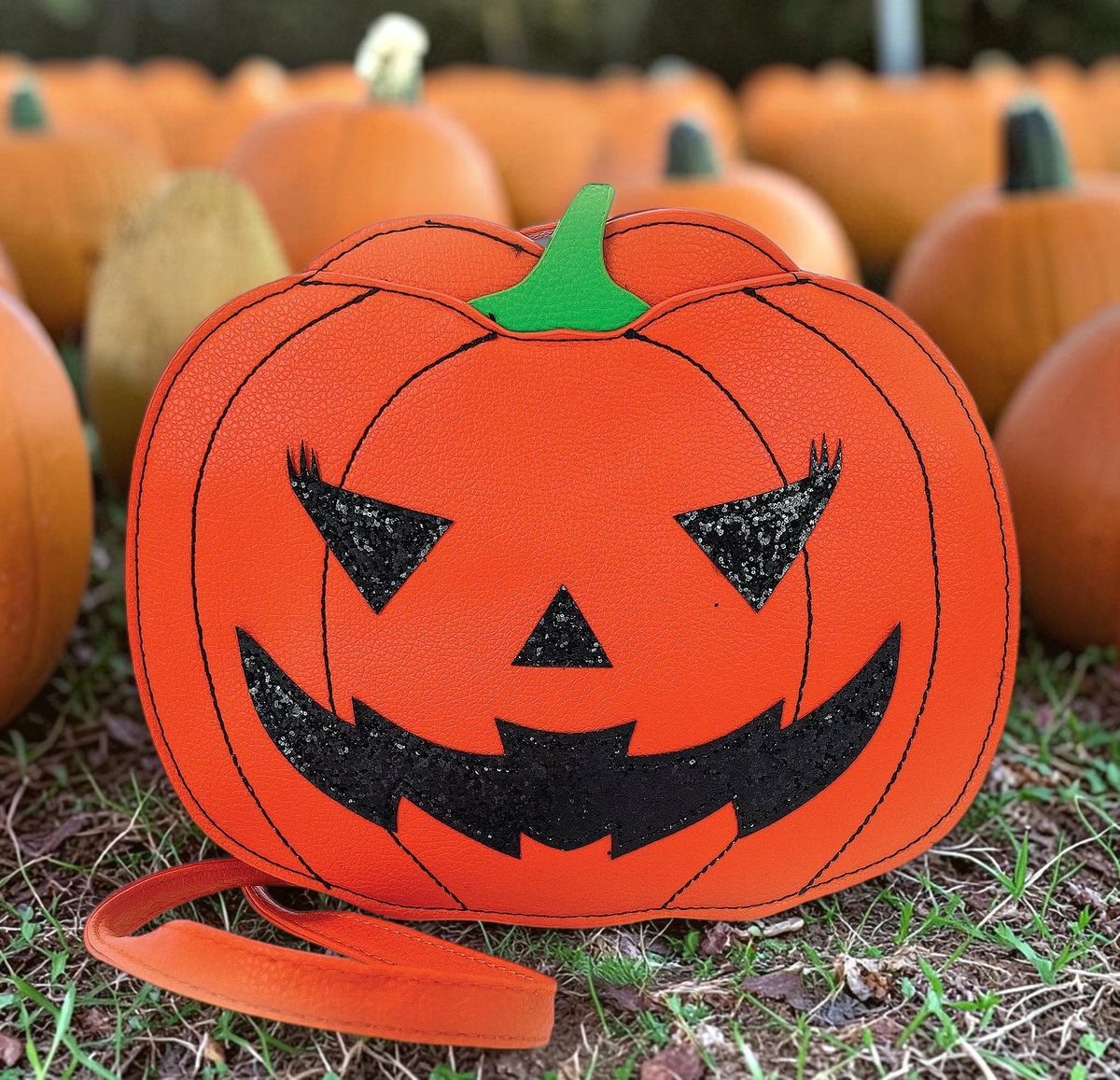 Halloween-Emporium orange vinyl jack-o-lantern crossbody bag with black glittery eyes and mouth in a pumpkin patch.