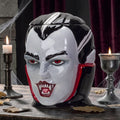 Halloween-Emporium vampire ceramic treat jar with glossy finish, red eyes, and fangs on a wooden table with candles.