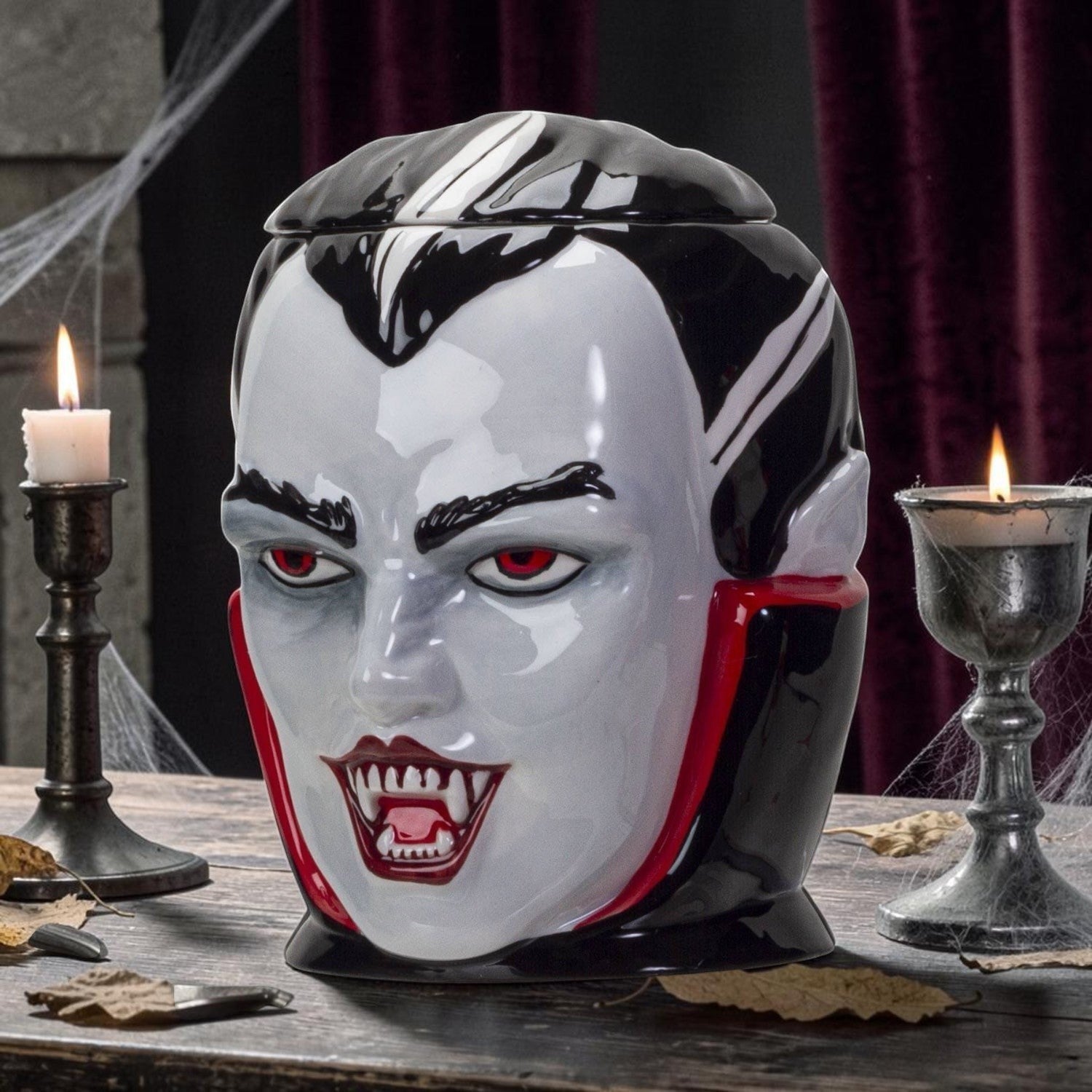 Halloween-Emporium vampire ceramic treat jar with glossy finish, red eyes, and fangs on a wooden table with candles.