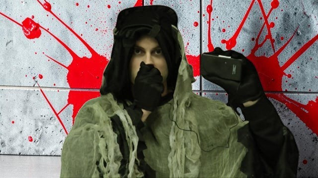 Halloween-Emporium voice changing accessory held by a person in a dark hooded costume against a blood-splattered wall