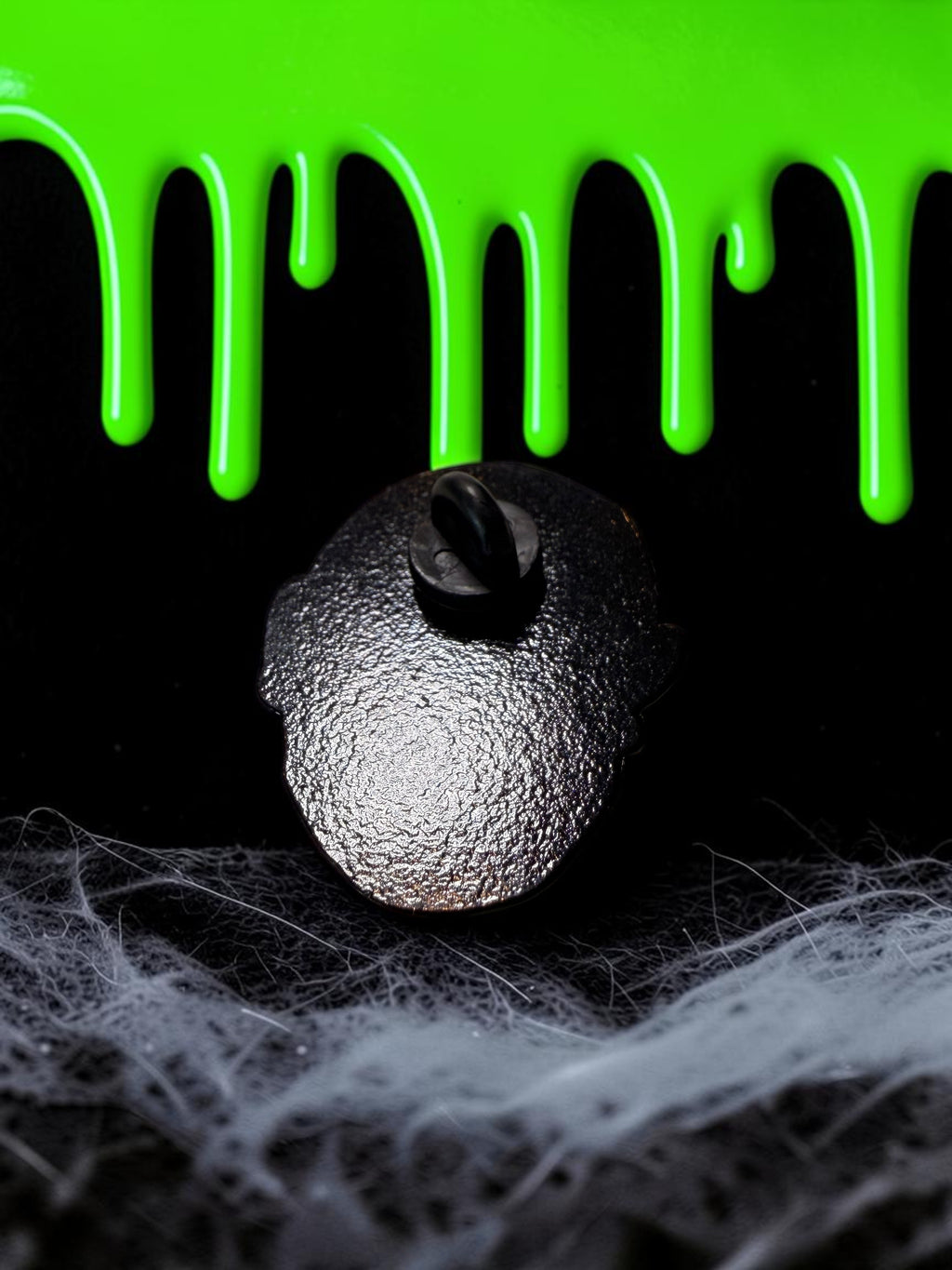 Halloween-Emporium zombie enamel pin back view with black metal finish against a green slime and spiderweb background